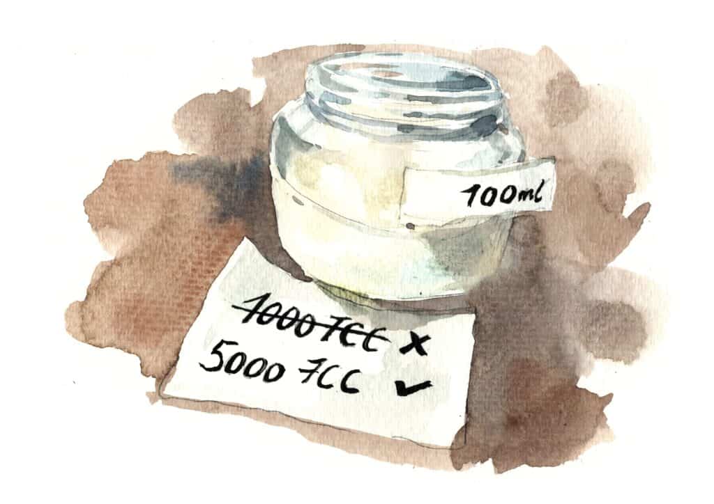 Glas Milch. Davor ein Zettel, auf dem die alte Laktase Dosierung von 1000 FCC pro 100ml durch 5000 FCC ersetzt wurde.